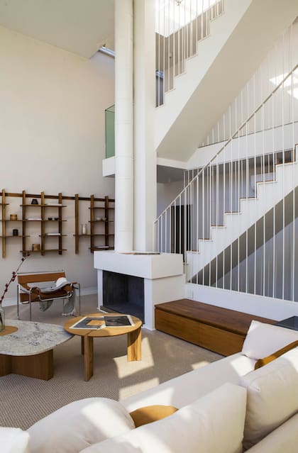 La chimenea como vértice de líneas funcionales que surgen de ella como rayos: el tiraje altísimo, un tramo de la escalera que conecta tres niveles y un mueble de asiento y guardado, vistos en una reforma de Estudio Plök.