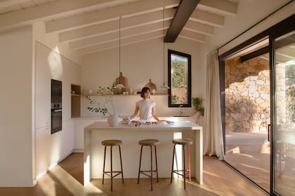 La cocina en L es funcional y liviana, con muebles blancos y estanterías de madera clara. La isla central, acompañada por taburetes de madera, introduce textura y calidez, rompiendo la monocromía sin perder armonía.