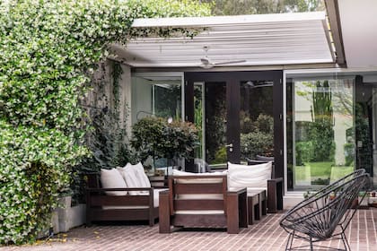 La galería con piso de adoquines de madera alberga un living con sillones cómodos para disfrutar del aroma del jazmín que crece por el muro y trepa por la pérgola