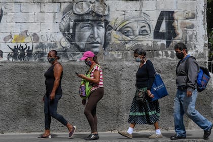 La gente usa máscaras faciales mientras camina junto a un mural que representa al difunto presidente de Venezuela, Hugo Chávez, en el centro de Caracas el 25 de agosto de 2020, en una semana en que el gobierno alivió un bloqueo nacional que se había impuesto para combatir la pandemia del nuevo coron