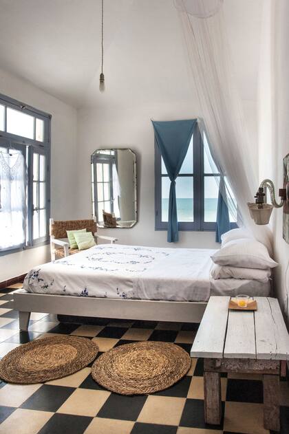 La mesa y la cama de líneas sencillas están acompañadas por alfombras de cardo, clásicas artesanías uruguayas. Los veladores y el espejo biselado se corren del estilo rústico y suman personalidad.