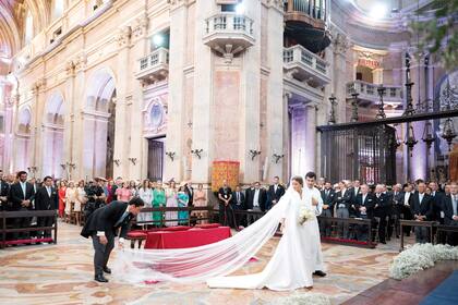La novia entra a la basílica y el novio le acomoda la cola del vestido, un diseño de Luzia do Nascimento confeccionado en seda mikado, con escote en V, hombros marcados y mangas francesas.