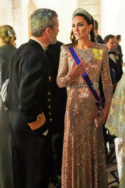 La princesa de Gales complementó su vestido de Jenny Packham con la tiara Lover’s Knot que en 1913 María de Teck,tatarabuela de William, encargó a la joyería Garrard. Le sumó la banda de la Real Orden Victoriana que le entregó Isabel II en 2017, la estrella de la Orden Real Victoriana y un broche art déco de diamantes.
