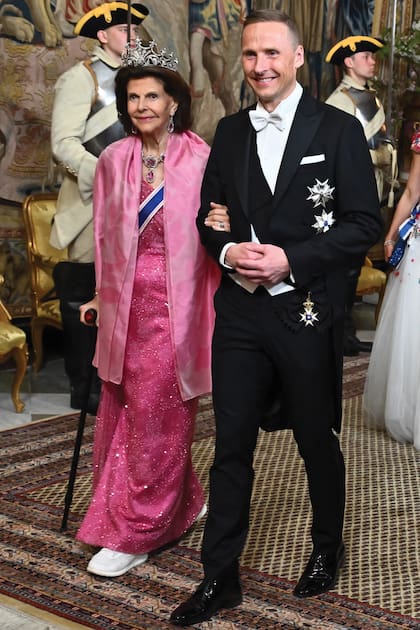 La reina Silvia entra al salón del Palacio Real de Estocolmo del brazo del marido de la presidenta de
Islandia, Björn Skúlason, y se ayuda
además con un bastón. Accesorizó su
traje con collar, pendientes y broches
de topacios rosados, la tiara de
nueve puntas y zapatillas deportivas
Tamaris.