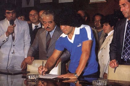 Maradona simula la firma de su contrato con Boca. Su garabato pareciera dibujarse en lo más alto de la hoja. A su lado, los presidentes de Boca, Martín Benito Noel, y de Argentinos, Próspero Cónsoli. Cerca suyo, como siempre, Cyterszpiler