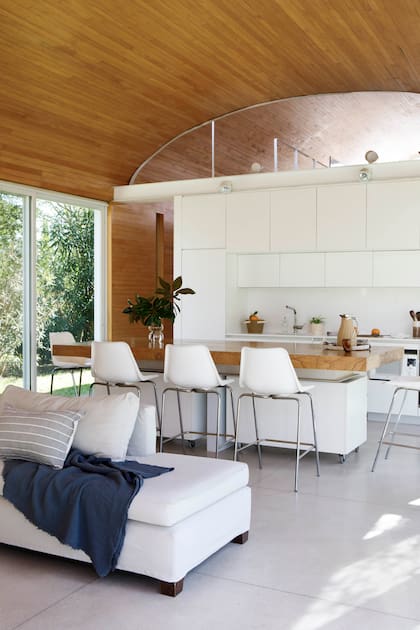 Mesa-isla con tapa de ciprés (Carpintería Sebastián Fernández Lando), banquetas (Hormiga Emergente). Recipiente de vidrio con arreglo verde, bandeja de fibras naturales, termo de ratán (Philippa Deco Boutique) y mate (Sasha Gats).