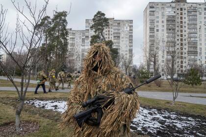 Miembros de las Fuerzas de Defensa Territorial de Ucrania pasan por el parque de la ciudad mientras un soldado camuflado se para en primer plano, en las afueras de Kiev, Ucrania, el miércoles 9 de marzo de 2022