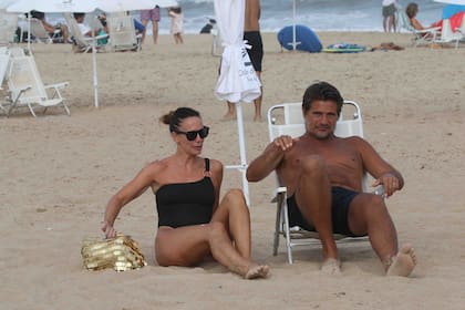 Mientras él viste un short de baño oscuro, ella marca tendencia con su enteriza, sus gafas y su bolso playero en dorado.