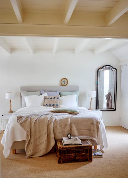 Moquete de fibras naturales, velador con base de tronco, baúl vintage, pie de cama y uno de los almohadones tejidos a mano: detalles que le dan un sofisticado toque rústico a un cuarto blanco.