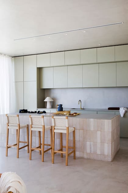Mueble de cocina (The Concrete Studio) con puertas ciegas, sin tiradores y con la heladera y el freezer panelados. Mesadas y alzada de placas cementicias. Grifería monocomando ‘Gold’ (Peirano).