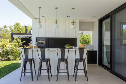 Parrilla con puerta guillotina de hierro negro (Metalfox). Revestimiento de siding blanco –el mismo de la fachada–. Barra con tapa de Purastone ‘Nube’ bajo tres lámparas de techo (Concreto Art). Un área de servicio, con mesada, bacha y espacio de guardado, hace de soporte cuando cocinan en la galería.