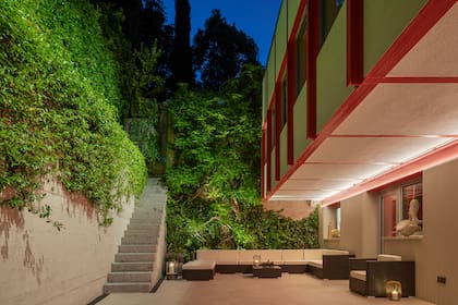 Patio lateral con la casa levitando a la derecha, sostenida por un exosqueleto estructural de vigas y columnas de acero. Largas franjas de luces LED circundan todos los costados de la propiedad.