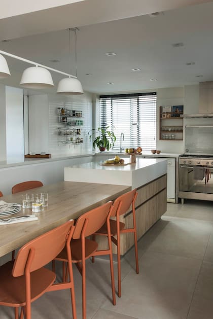 Piso de porcelanato (SBG) y amoblamiento (Arquimadera) en laca blanca con mesada en Silestone. La isla central baja su altura y se prolonga en una mesa de comedor diario con revestimiento símil madera. Luminarias (Via Luce). Sillas de hierro y madera color ladrillo (El Yeite).