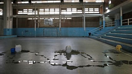 Polideportivo Chacabuco. Uno de los gimnasios internos tiene varias filtraciones en el techo; en un día de lluvia, las goteras invaden la cancha; al fondo se observan los vidrios rotos