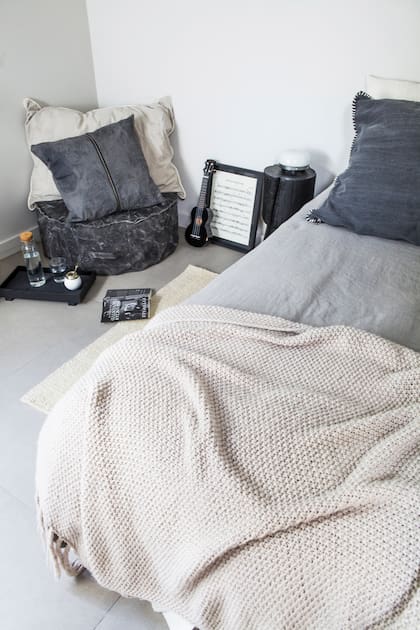 Ropa de cama y cover, almohadón ‘Santa’, almohadón con cierre y genero engomado, mesa de madera natural con lustre negro, puf de lona gris teñida artesanalmente (todo de Paul French Gallery). Alfombra de lana natural (Elementos Argentinos).