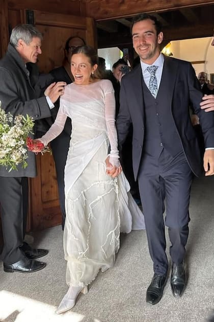 Santi y Maggie, recién casados, en la puerta de la capilla. Ella eligió un vestido de tul con mangas largas, sobre un viso en color blanco roto y botitas blancas de raso en punta. Llevó, además, un ramo de estilo silvestre de flores blancas y el pelo semirrecogido, peinado con ondas naturales.