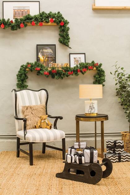 Sillón vintage rayado, mesa auxiliar de paraíso con dorado y lámpara de conejos frente a un trineo de madera. Los estantes flotantes se recubrieron con muérdagos y guirnaldas de luces, con buses londinenses (Salazar Casa)