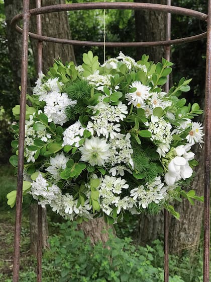 Sobre una base de oasis armada con alambre para poder colgar, este arreglo tiene una estructura verde de buxus y asparagus y flores blancas de verbenas, nigelias, orlayas, rosas y aster.