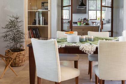 Vista hacia la cocina desde la mesa con un mantel de hilo (Madison Market). En el rincón, canasto con palmera (Soleil Deco) y banquito de cuero (Laro).