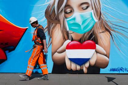 Un albañil pasa frente a un mural del artista Casper Cruse de una mujer con mascarilla sosteniendo un corazón con los colores de la bandera holandesa en apoyo a quienes sufren por los efectos de coronavirus en La Haya, Holanda, el viernes 24 de abril de 2020. (AP Foto/Peter Dejong)