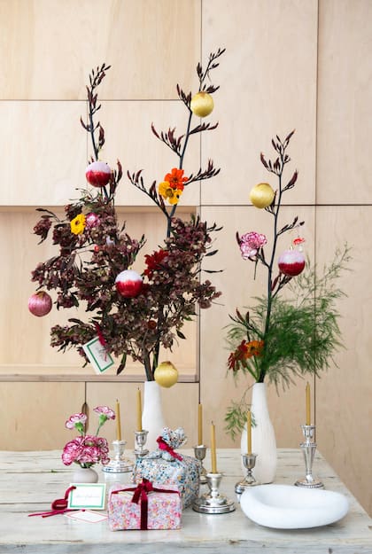 Un arreglo floral también puede cumplir la función de arbolito de Navidad. En este caso, varas de formios, follaje verde y violáceo, zinias y el toque especial: bolas o esferas que aportan color y aire festivo. También, un florero con claveles, candelabros de diferentes tamaños y regalos envueltos en género con cintas de terciopelo y tarjetas con los nombres.