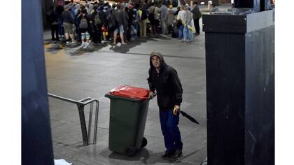 Un hombre empuja un tacho de basura, detrás un grupo de personas espera recibir comida en el refugio del centro de Sidney