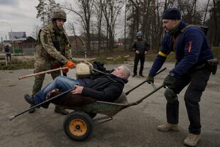Un hombre es trasladado en una carretilla tras cruzar un río por un camino improvisado bajo un puente destruido por un ataque aéreo ruso, mientras la gente huye de la localidad de Irpin, Ucrania, el sábado 5 de marzo de 2022. (AP Foto/Vadim Ghirda)