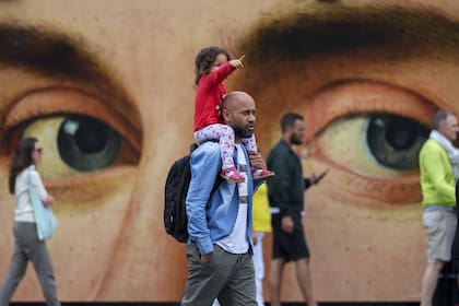 Un hombre lleva a una niña sobre sus hombros, caminando junto a un cartel que muestra una pintura del artista italiano Antonello da Messina afuera de la Galería Nacional en Trafalgar Square de Londres, el lunes 1 de julio de 2024.