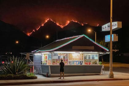 Un hombre se encuentra frente a un restaurante de comida para llevar mientras las llamas arden en las colinas del Bosque Nacional de Ángeles en el fondo en medio de los incendios forestales en California en septiembre de 2020.