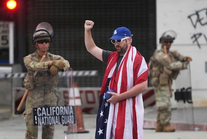 Un manifestante cubierto con la bandera de Estados Unidos alza un brazo frente al Centro de Detención Metropolitano y miembros de la Guardia Nacional de California, el jueves 12 de junio de 2025, en Los Ángeles