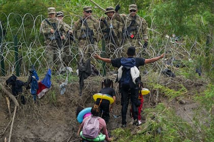 Un migrante abre los brazos frente a miembros de la Guardia Nacional de Texas ubicados detrás de alambre de púas en una de las márgenes del río Bravo, el jueves 11 de mayo de 2023, vistos desde Matamoros, México (AP Foto/Fernando Llano)