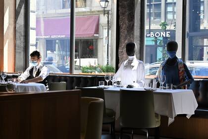Un mozo con una máscara facial ajusta las copas en una mesa junto a los maniquíes colocados para proporcionar distancia social en un restaurante en el viejo Montreal el 10 de julio de 2020 en medio de la pandemia de coronavirus