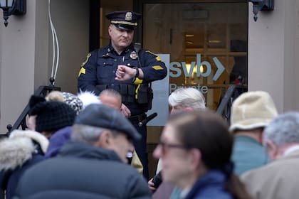 Un oficial de policía de Wellesley, Massachusetts, mira su reloj mientras los clientes y transeúntes forman una fila fuera de una sucursal del Silicon Valley Bank, el lunes 13 de marzo de 2023.