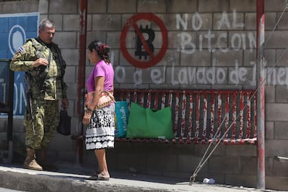 Un soldado habla con una vecina en una parada de ómnibus cubierta con graffiti en contra del uso del Bitcoin, antes de unas elecciones municipales en San José Las Flores, El Salvador