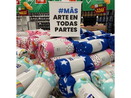 Una escena evoca las fotografías de la serie Pop Latino de Marcos López: un cubo de cartón anuncia el título del programa, “Mâs arte en todas partes” sobre una pila de mantas polares en venta