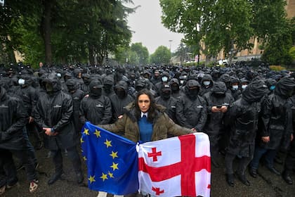 Una mujer sostiene una bandera de Georgia y una de la UE frente a agentes de la policía que impiden el paso a los manifestantes durante una protesta contra una ley cerca del Parlamento en el centro de Tiflis, Georgia, el martes 14 de mayo de 2024. (AP Foto/Zurab Tsertsvadze)