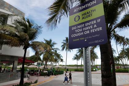 Una pareja pasa junto a un cartel que pide a las personas que no visiten Miami Beach, la famosa South Beach de Florida si tienen tos o fiebre, el lunes 22 de junio de 2020. Más de 100,000 personas en Florida han sido diagnosticadas con el coronavirus, informaron funcionarios de salud estatales.