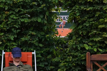 Una tenista ejecuta un golpe a media pista en una pista de tierra batida en Roland Garros, vista a través de un seto con un espectador descansando al fondo
