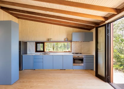 La biblioteca se prolonga para ocultar la heladera y anticipa el amoblamiento de cocina laqueado en el mismo tono. Una ventana apaisada sobre la mesada de mármol de Carrara apunta directamente al pinar.