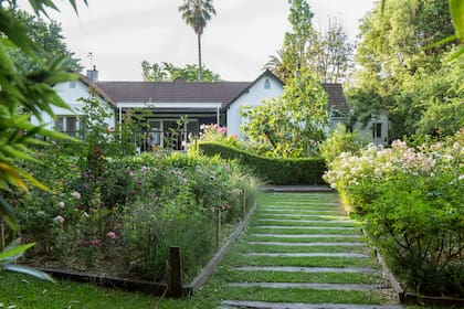 Vista a la casa que sale del muelle con un camino de durmientes, canteros de rosas, un cerco de buxus y detrás una higuera, achiras y más rosas.