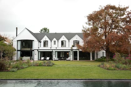 Vista general desde la pileta. Grandes macizos coloridos se recortan contra el césped y enmarcan la clásica construcción puesta en valor con colores neutros. El añoso taxodium se destaca como el gran protagonista.